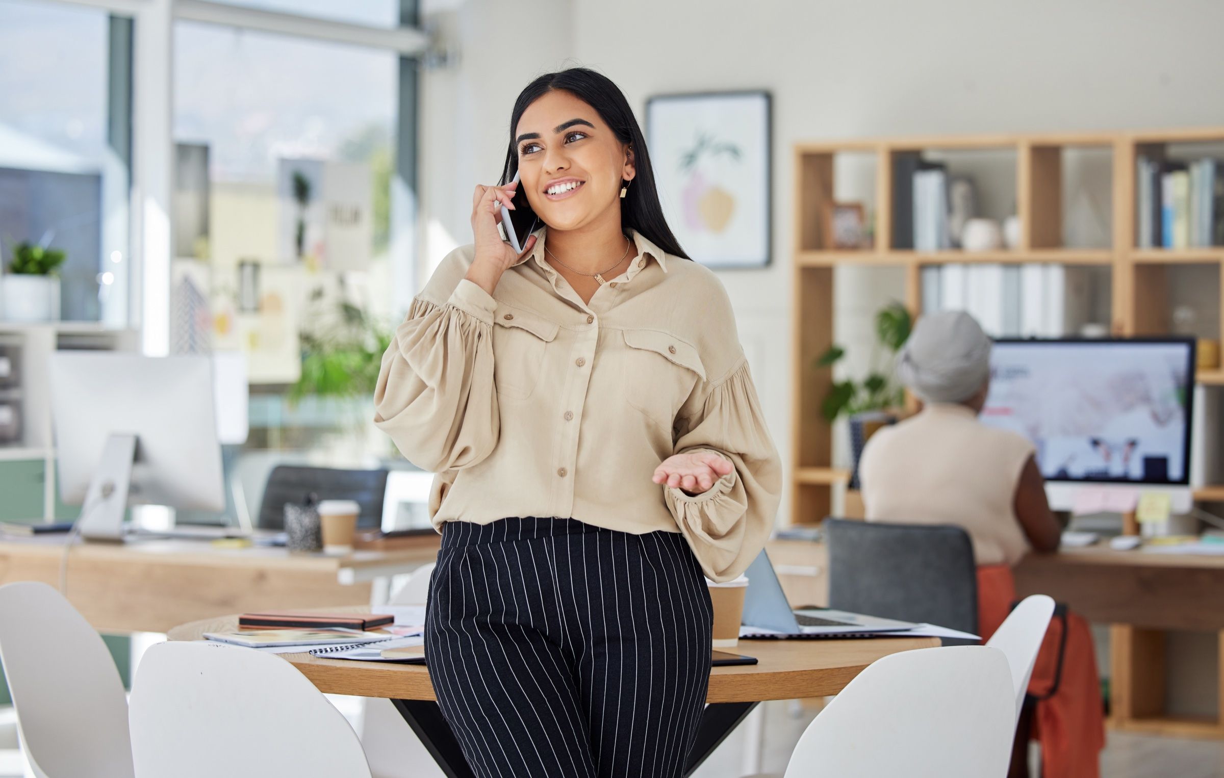 Mujer sonriendo mientras habla por teléfono en una oficina, de pie junto a una mesa de trabajo.